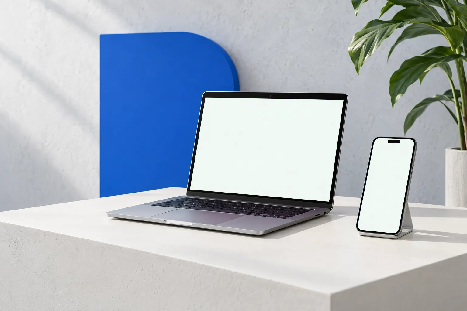 Portátil y teléfono sobre una mesa clara, junto a un panel azul y una planta, con un sitio web de ejemplo en pantalla